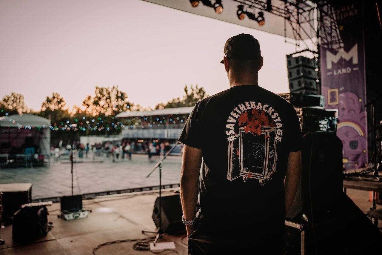 Zsolt seen from behind on stage at dusk, wearing a #SaveTheBackstage T-shirt
